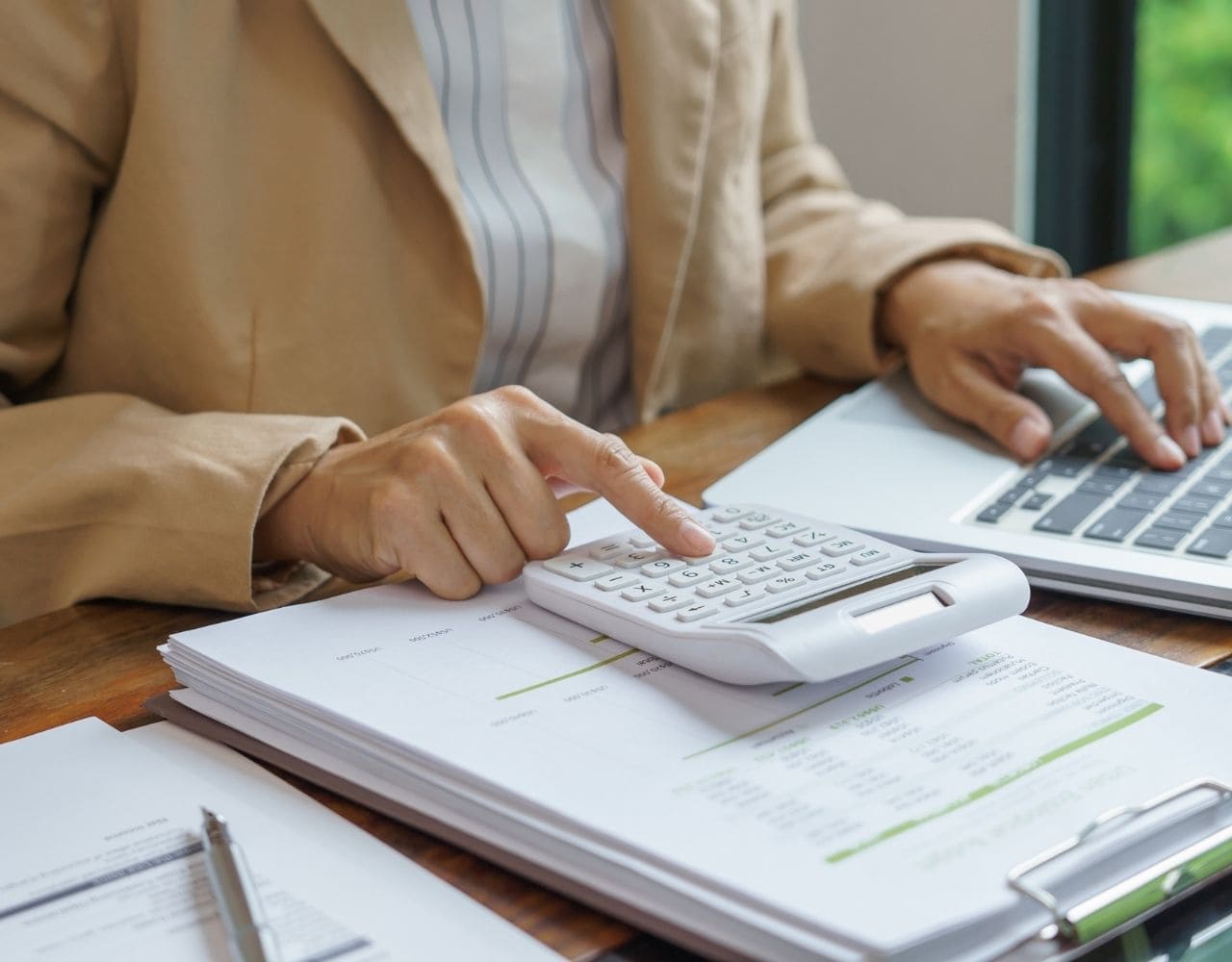 Une personne concentrée effectue des calculs sur une calculatrice, des documents financiers et un ordinateur portable devant elle. L'image représente la préparation d'une liasse fiscale.